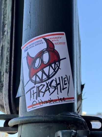 A hand drawn sticker by Thrashley. It shows a demon's head with jagged teeth and horns. It is drawn in a cartoony style with red and black ink on a USPS sticker. Below the demon is text that says "Thrashley @thrash_lee".