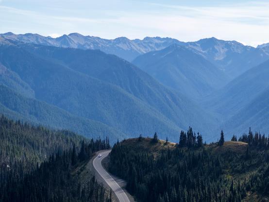 A winding road cuts through dense evergreen forests in a mountainous landscape, with rolling peaks and a clear blue sky in the background. A lone car travels along the road, emphasizing the remote beauty and vastness of the natural scenery.