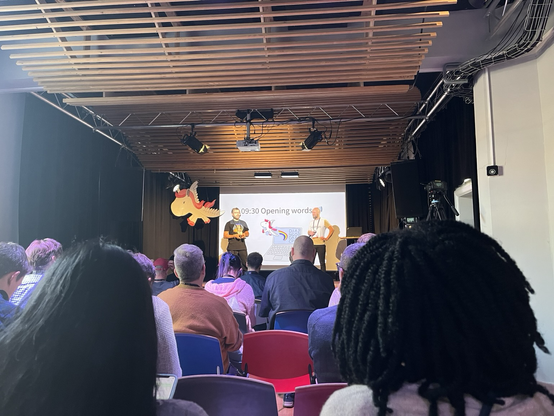 A stage with a slide saying “Opening words”. Organizers Ben and Emil are in front. A wooden Django pony hangs from the ceiling.