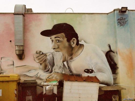 Farbfoto eines Straßenkunstgemäldes an einer Wand in Torvaianica, Lazio, Italien. Es ist eine Farbkopie einer bekannten Szene aus dem italienischen Schwarzweißfilm Ein Amerikaner in Rom von 1954 und zeigt einen Mann mit Schirmmütze beim Verschlingen einer Riesenmenge Spaghetti. Leica R4 / 35 mm Summicron / Ilford Ilfocolor 400