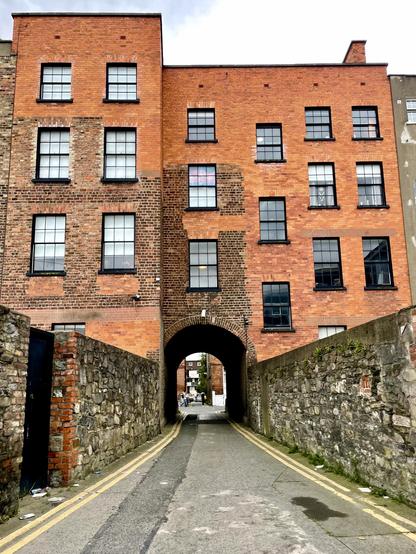 A brick building with large windows features an archway at the base, leading into a narrow alleyway lined with stone walls. The scene is partially cloudy, with visible pavement and greenery in the background.