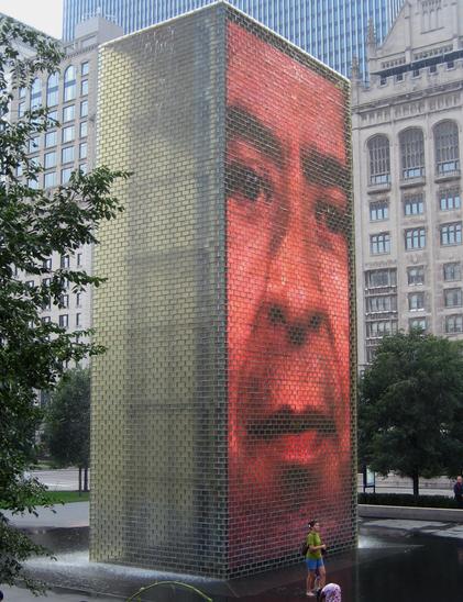 A large, illuminated brick structure in a public space features a close-up image of a person's face, partially obscured by water. People are seen near the base, with greenery and urban buildings in the background.