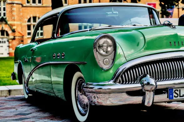 Ein Auto der Marke Buick aus den fünfziger Jahren ist von der rechten Seite zu sehen. Die Front zeigt viel Chrom und elegante Linien. Das Auto ist grün mit einem weißen Dach. Die Weißwandreifen leuchten in der Sonne. Im Hintergrund das Gebäude des Museumsberges in Flensburg. 
.
A Buick car from the 1950s can be seen on his right. The front shows a lot of chrome and elegant lines. The car is green with a white roof. The whitewall tyres glow in the sun. The Museum Hill building in Flensburg is in the background. 
.