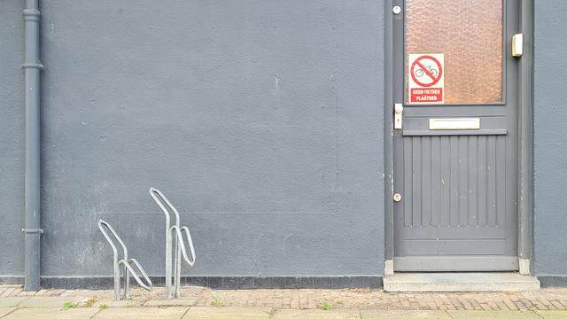 Two empty bike stands on a sidewalk in front a grey painted concrete wall with a similarly painted drainpipe. A meter to the right of the stands a slightly darter grey, wooden door with a mail slot, locks at the top, middle and bottom of the left side and a window. In the bottom left of the window a large rectangular sticker with a no parking bicycles sign above the text do not place bikes in white capital letters in a red rectangular box.