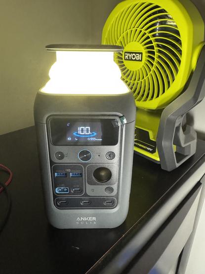 An Anker SOLIX C300 DC Power Station is sitting on a nightstand with the built-in light extended. Behind it is a Ryobi fan which runs on their 18 volt batteries.