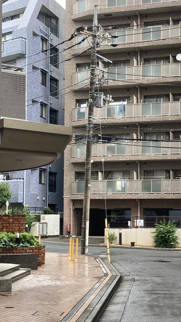 A street view featuring a tilted utility pole with wires, adjacent to a modern apartment building. The foreground includes a brick planter and a paved walkway leading to a corner of the street.