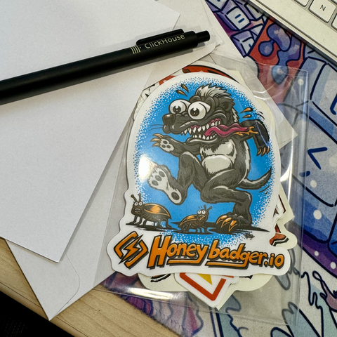 A desk scene with a black pen labeled "ClickHouse" on a piece of paper. Next to it is a colorful sticker featuring a cartoon honey badger character with big eyes and sharp teeth. The sticker has "Honeybadger.io" written at the bottom. In the background, part of a keyboard and some other papers or stickers are visible.