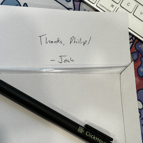 A close-up of a handwritten note on a piece of paper. The note reads "Thanks, Philip! - Josh". Below the note, part of a transparent plastic envelope or sleeve is visible. A black pen with "ClickHouse" written on it is partially shown at the bottom of the image. The edge of a keyboard and some colorful designs (possibly stickers) can be seen in the background.