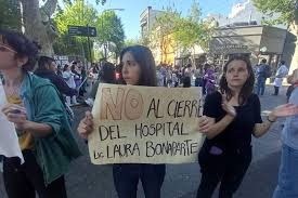 Foto de la protesta por el cierre del Hospital Bonaparte en la intersección de la calle Combate de los Pozos y Avenida Caseros. Se ven a dos personas sosteniendo carteles. Uno de ellos dice: "NO al cierre del Hospital Lic. Laura Bonaparte". Se ve a otra persona aplaudiendo.