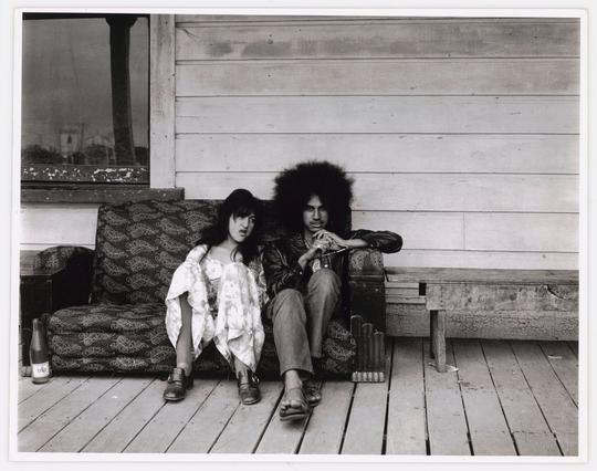 The image is a black and white photograph depicting two individuals sitting on an outdoor bench. The person on the left appears to be wearing casual attire with layered clothing, including what looks like a kimono-style garment over another outfit. Their hair is short and they have glasses. Next to them sits someone whose features are more pronounced due to their larger afro hairstyle. This individual wears dark-colored jacket or coat sleeves that extend down past the wrist and has a relaxed posture.

In front of this person, on what seems like a wooden deck or porch area, there's an assortment of objects including bottles with labels partially visible in the background which could suggest casual drinking activity. The bench they are seated upon is worn and shows signs of use such as faded paint and wear marks from weather exposure.

The setting suggests a rural or countryside environment, potentially outside someone’s residence given the presence of personal items like suitcases beside them. There's also an old-fashioned bottle with visible labeling next to one individual which adds context suggesting they might have been enjoying beverages in this casual outdoor space. The backdrop is quite simple and nondescript but consists mainly of a wooden structure, likely part of their home or another dwelling.

The photograph captures the essence of everyday life and human conne [...]