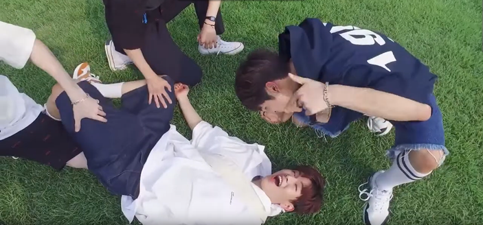 Seungmin lies on the grass in a park. Two people have their hands on his thighs, and a third person is crouched over his face and singing down at him. Seungmin is very happy.