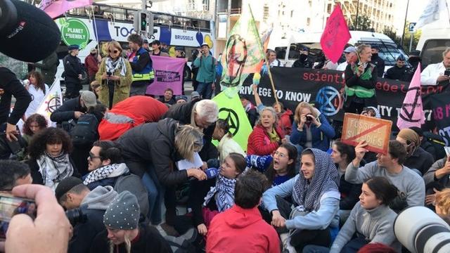 Greta Thunberg among a crowd of activist sitting in a road in Brussels. There are Scientist Rebellion and Extinction Rebellion banners and flags as well as symbols of Palestinian solidarity.