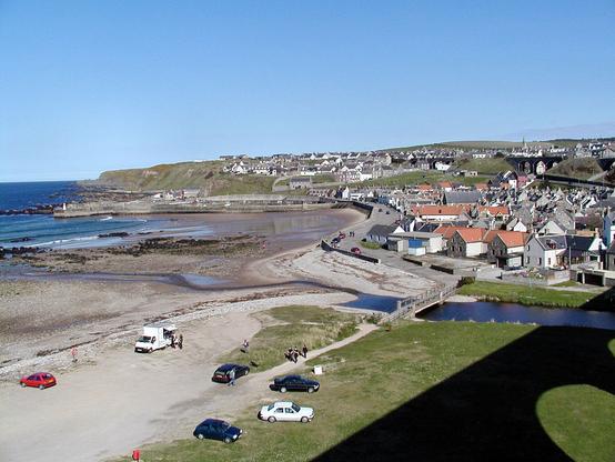 Cullen. The image shows a view from an old railway viaduct, whose shadow is in the bottom-right, over a broad beach in the lower left of the frame to the blue sea on the left. A headland rises beyond the bay and the town extends from beyond a river on the right around to the headland. The sky is light blue.