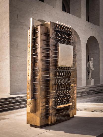 Scultura d’arte moderna a forma di parallelepipedo. Presenta varie forme geometriche e sporgenze sulla sua superficie, conferendole un aspetto complesso e strutturato. La scultura è realizzata in un materiale metallico con una finitura riflettente e dorata. È esposta all’aperto davanti a un edificio con archi in pietra e una statua nelle vicinanze, creando un ambiente artistico contemporaneo