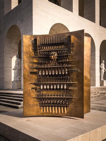 Scultura d’arte moderna a forma di parallelepipedo. Presenta varie forme geometriche e sporgenze sulla sua superficie, conferendole un aspetto complesso e strutturato. La scultura è realizzata in un materiale metallico con una finitura riflettente e dorata. È esposta all’aperto davanti a un edificio con archi in pietra e una statua nelle vicinanze, creando un ambiente artistico contemporaneo
