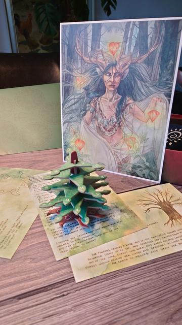 Eine aufrecht stehende Grafik einer mystischen Frauenfigur mit Geweih und wehendem Haar, die an Faden kleine herzförmige Laternen hält. Davor ein kleiner aus Pappteilen zusammengesteckter Baum