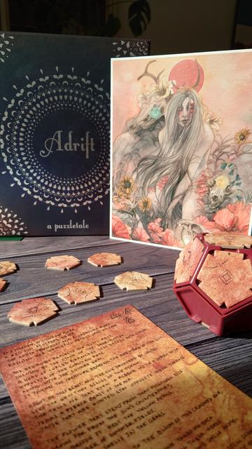 Vor dem Karton in abstrakten Design mit der Aufschrift Adrift steht aufrecht eine Grafik einer mythischen Frauenfigur mit langem Haar. Sie ist umgeben von Blumen, Edelsteinen und Tierschädeln.

Davor fünfeckige Pappteile, ein Dodekaeder aus Plastik un dessen Seiten ein paar der Pappteile eingesetzt wurden und ein Gedicht, alles in rot und Brauntönen gehalten