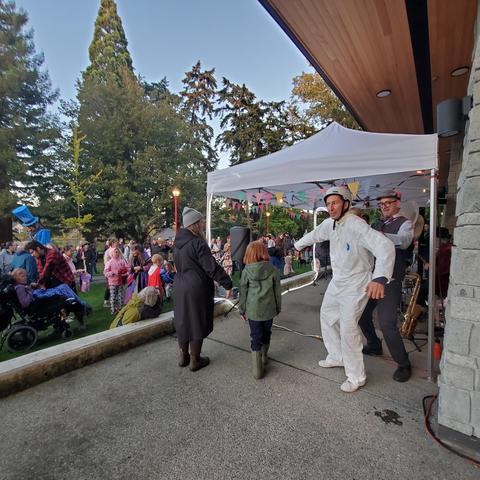 A man in all white long sleeve coveralls with white hard helmet getting zipped up in back by musician under performance stage tent. A small crowd is gathering on the left.