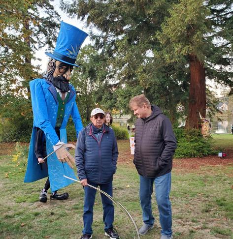A giant walking puppet, bearded man with blue tophat & blue long tuxedo jacket is lurking behind two men chatting. A young female volunteer is seen behind the men, being walking puppet safety/security.