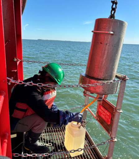 A technician on a ship sampling stage collects water from a large metal container. The water is green and islands are seen on the horizon.