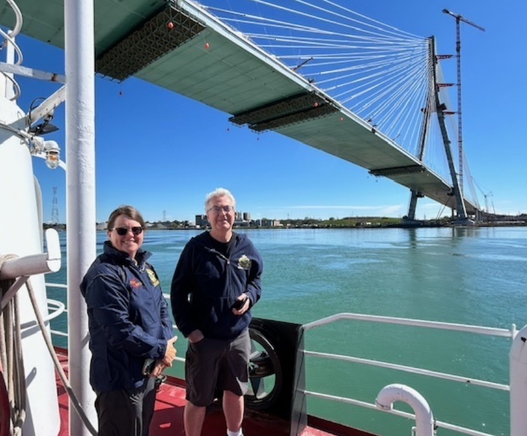 Two Fisheries and Oceans Canada science staff are standing on the deck of ship passing underneath a large suspension bridge still under construction.