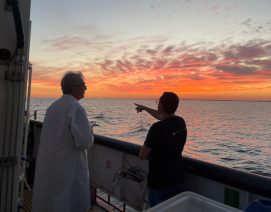 Under a dark orange sunset over a large lake, one of a pair of science crew members on a ship points in the direction of the horizon.