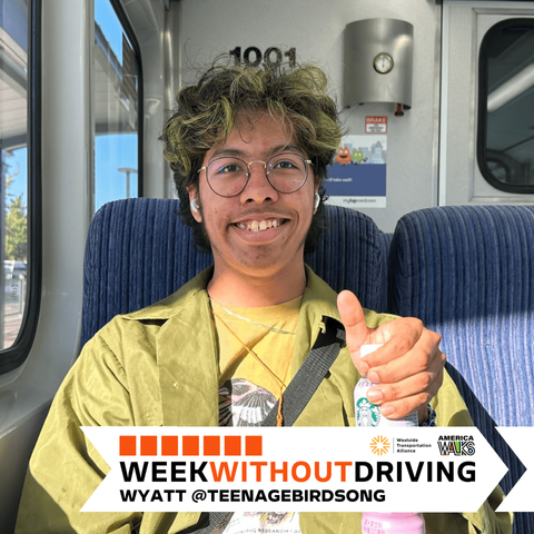 Wyatt, sitting in a WES train, wearing yellow shirt, and a green jacket. Holding up a drink bottle and giving a thumbs up. Week Without Driving Banner, Westside Transportation Alliance and America Walks logos, "Week Without Driving, Wyatt @TEENAGEBIRDSONG"