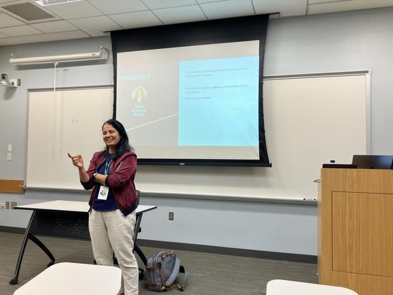 An individual is presenting at a conference or workshop, standing in front of a projector screen displaying a "Thank You!" slide. The person, identified as Mansi, is smiling and gesturing, wearing a maroon jacket over a blue shirt.