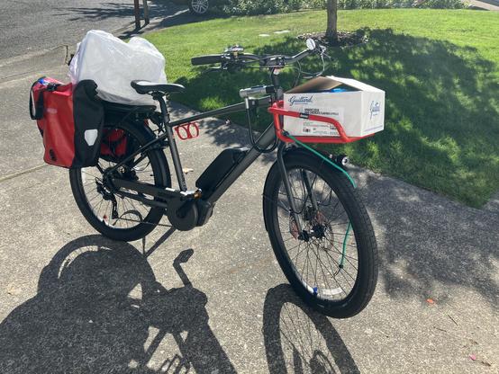 A dark grey electric bicycle with red panniers and a red front rack. The front rack has a large white box in it and the rear rack has a larger white box covered with plastic.