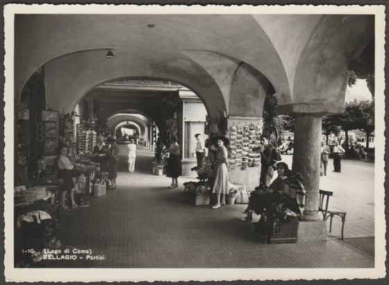 Monochrome real photographic postcard showing a view of the Portici in Bellagio on Lake Como in Lombardy, Italy, and showing a rack of postcards for sale.
Published by S. Lanfranconi, Bellagio, No 1-10, c.1950s.
Postally unused.
Very good condition, with very slight corner bumps.