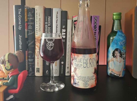 A 5oz stemmed glass filled with beer and a 500ml bottle sit on a shelf. The glass has the Sapwood Cellars logo (half hop/half acorn) w/ antlers on top. Behind on the shelf are various books on music and music artists, and a soju bottle featuring Hyori. A LoFi Girl figure is to the left.