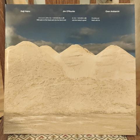 LP cover. Photo of mountains of sand, or sand dunes, with blue sky and white clouds in the background. 

In white text on top: Keiji Haino / Jim O'Rourke / Oren Ambarchi
"With pats on the head, just one too few is evil one too many is good that's all it is"