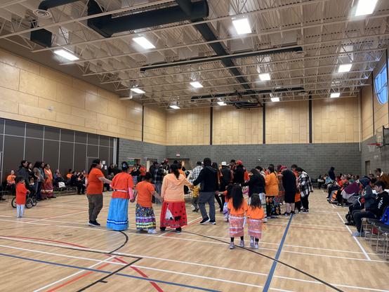 People, including those dressed in orange shirts and ribbon skirts, taking part in a Round Dance.