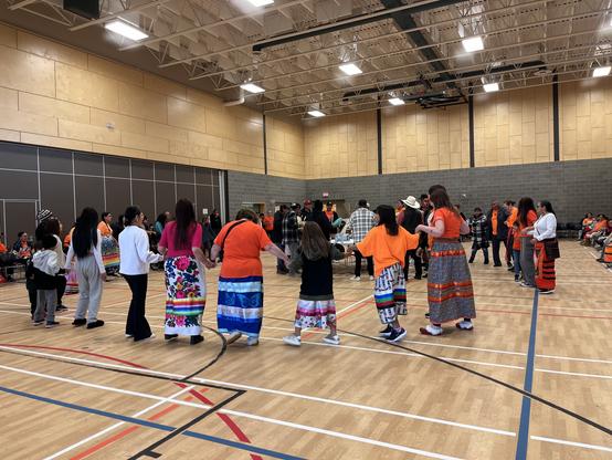 People, including those dressed in orange shirts and ribbon skirts, taking part in a Round Dance.