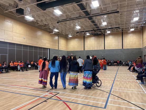 People, including those dressed in orange shirts and ribbon skirts, taking part in a Round Dance.