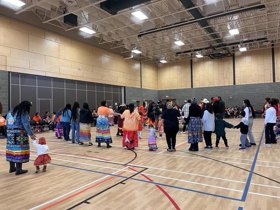 People, including those dressed in orange shirts and ribbon skirts, taking part in a Round Dance.