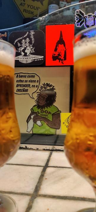 En primer plano se ven un par de cervezas algo desenfocadas. Al fondo se ven unas pegatinas en un metacrilato de diferentes grupos andaluces. En el centro de la imagen hay un dibujo de un mono con una camiseta verde del bar loop que limpia un vaso. Del mono sale un bocadillo en el que se puede leer: "A bares como estos se viene a aprender, no a enseñar".