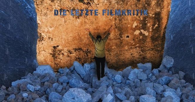 Visuell faszinierende Darstellung eines Megalithen im Film Architecton. Der Megalith ist hell orange und er sticht so aus dem ansonsten in dunkelblaue Farbtöne gehüllten Rest des Bildes hervor. Davor steht der weltbekannte Architekt Michele De Lucchi mit dem Rücken zu uns und streckt seine Arme in die Höhe, mit seinen Händen an den Megalith gelehnt. Coverbild des Podcasts "Die Letzte Filmkritik".