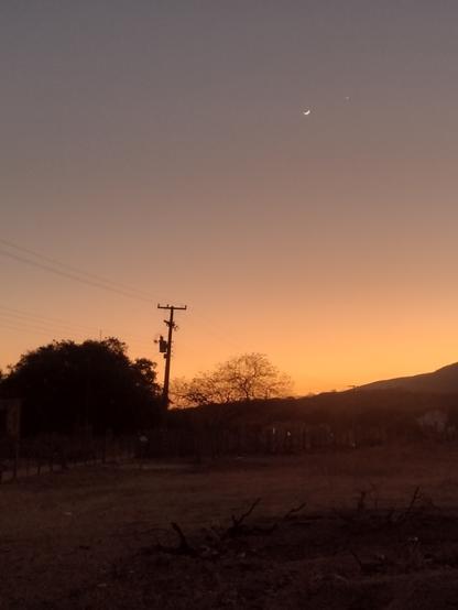 Foto de um por do sol na roça. A foto tem um contraste entre o céu laranja e o resto do cenário escuro quase preto. Dá para ver a silhueta de um poste, da serra ao fundo, uma árvore. No céu dá para ver a lua minguante e uma estrela.