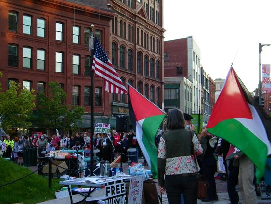A photograph of a rally in a city with brick buildings from the 1880s. There are many Red, Green, White and Black Palestinian flags, as well as signs that read, "End the Occupation", "Free Palestine" and "Maine Voices for Peace."