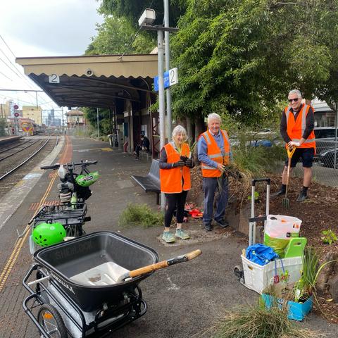 Volunteers in hi-vis vests pose with gardening tools on a train platform garden