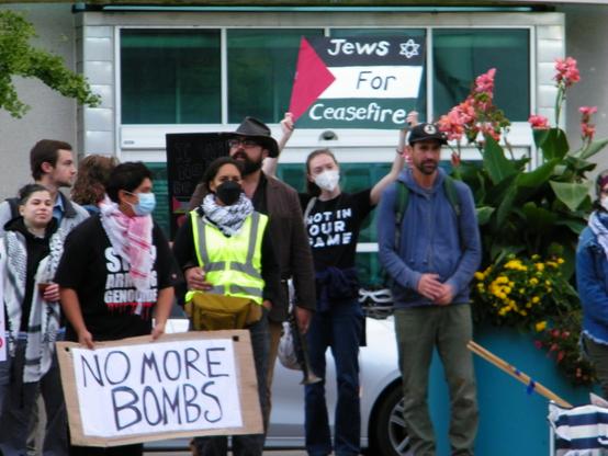 Protestors at an event. Two figures are promintent. One is wearing a t-shirt with ""Stop Arming Genocide" and holding a sign that reads, NO MORE BOMBS. Another is wearing a t-shirt with "Not In Our Name" and holding a Palestinian flag with the words "Jew For Ceasefire" in the stripes.