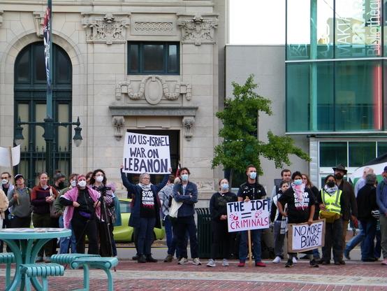 Protestors standing in front of an old building next to a modern one.
One of  the protestors is holding a sign that reads, "No US-Israel War on Lebanon." Another holds a sign that reads, "The IDF = Criminals".