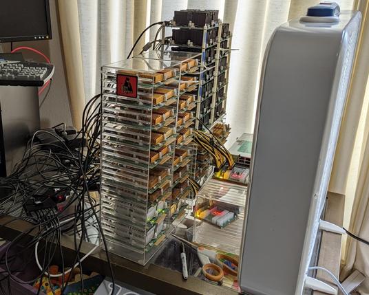 A stack of 36 ZTEX FPGA boards in a couple of acrylic CD towers, fed through a rat's nest of USB hubs. A box fan sits in front. A John the Ripper sticker graces the edge of the outer stack. Some heatsinks are copper-colored and less than an inch high; others are black and taller.