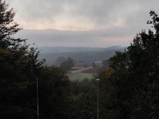 Photo of a valley through trees. There is morning mist. Several "layers" of hills are under several layers of clouds. Above the clouds the brush blue sky is an early morning is visible and there are reddish hints of the sun