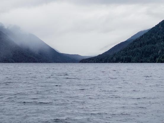 A serene, wide lake with gentle ripples stretches between two forested mountain ranges. The sky is overcast, creating a calm, misty atmosphere.