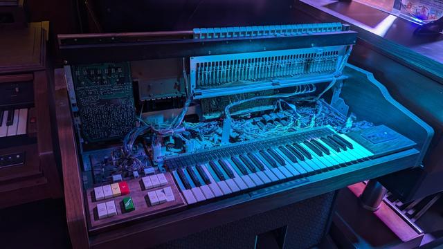 photo of an opened early electronic keyboard music instrument. it's illuminated with blue light. one can see the keys, switches as well as all the wiring.