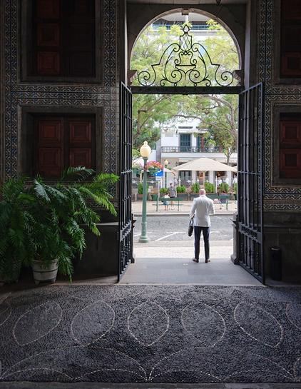 A man in a white jacket stands in a large gateway facing outwards. The foreground is quite dark but decorative floor mosaic and painted wall tiles are visible surrounding the gate and a large potted plant is highlighted by the light coming in through the gate. Outside the gate we can see a brightly sunlit street with decorative lamppost, trees, umbrellas and terraces as well as people sitting on benches.