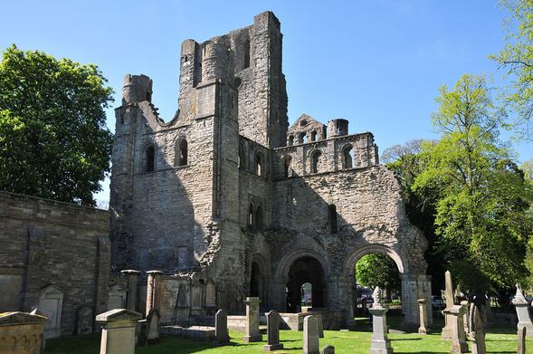 The ruins of Kelso Abbey. The image shows a diagonal view of the grey stone ruin of part of the abbey church at Kelso, standing in one place to the height of the tower with lower segments to its left and right. In the foreground, what would once have been the interior of the abbey church is grassed over and covered with gravestones. There are trees both sides of the ruin. The sky is blue.