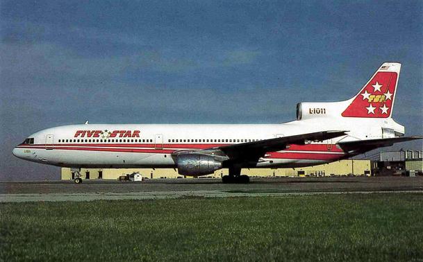 Still wearing its TWA livery, this plane has its new airline name applied over. It's parked on a ramp under gray skies.